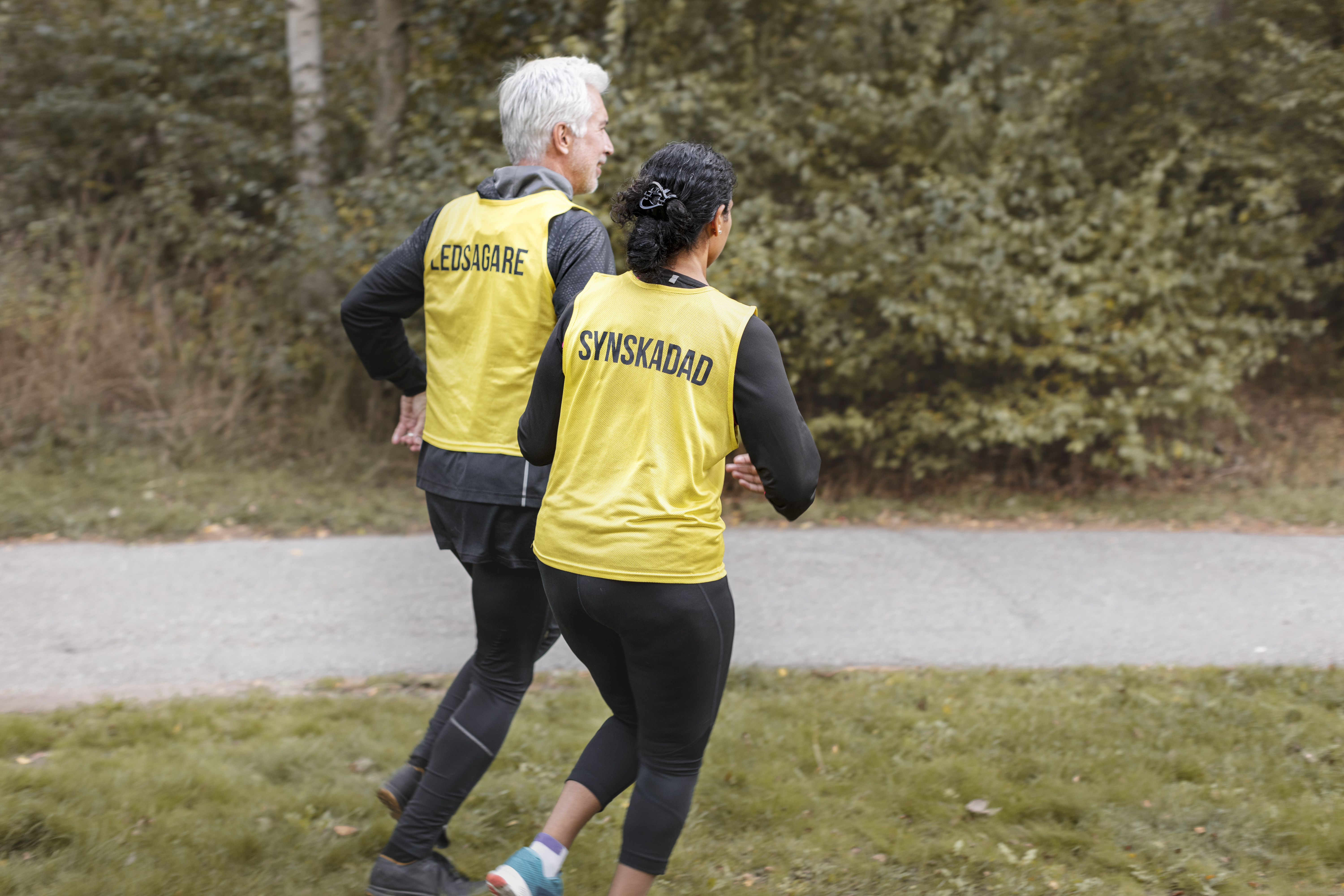 Två personer syns i helfigur bakifrån, de joggar tillsammans vid ett skogsområde. Mannen har grått hår och en väst det står "Ledsagare". Kvinnan har svart hår i hästsvans och en väst det det står "Synskadad".