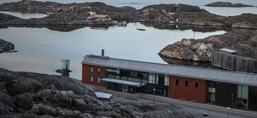 Ett kustlandskap med klippor och lugnt vatten, där Nordiska Akvarellmuseet ligger i förgrunden som en modern byggnad vid havet. I bakgrunden syns små öar och bebyggelse som speglar sig i vattnet.