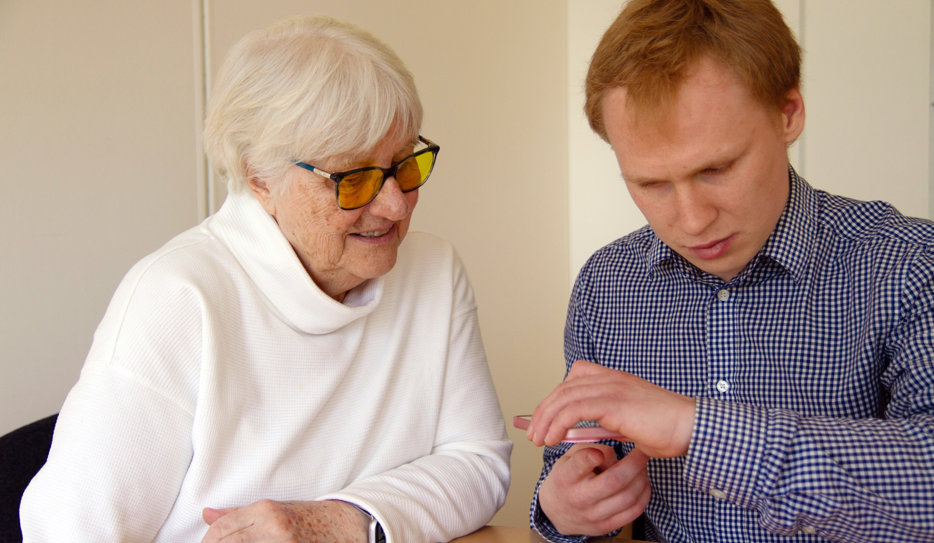 Äldre kvinna och yngre man vid sitter bredvid varandra. Han visar något på sin telefon och hon tittar intresserat.