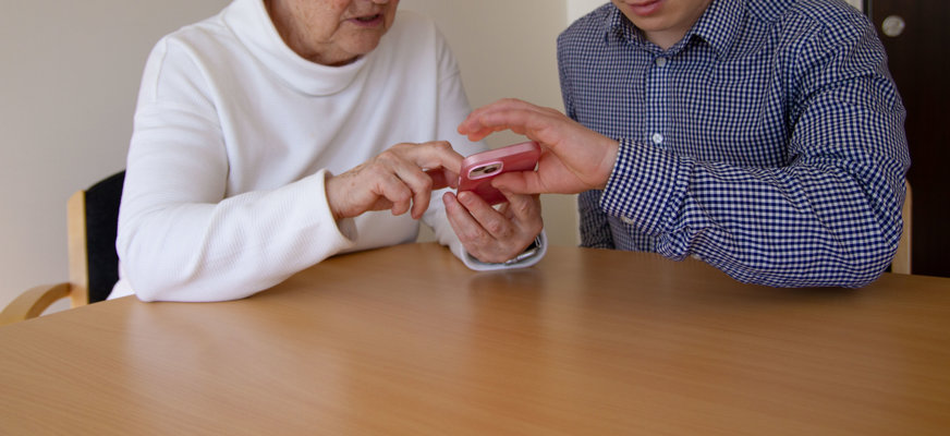 En yngre person visar en äldre person hur man använder en mobiltelefon.