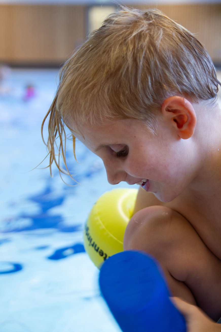 Närbild barn i profil, blött hår, armpuff, i bakgrunden simbassäng.