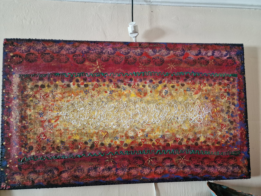 **Syntolkning (max 1000 tecken):**  Ett färgstarkt abstrakt konstverk med en central yta fylld av virvlande, vit skriptliknande text som liknar handskrift men inte bildar igenkännbara ord. Runt denna mitt breder sig varma färger – gult, orange och rött – med små cirkulära och blomliknande motiv. Ytterkanterna är mörkröda och lila, dekorerade med upprepade cirkelmönster och gröna sicksacklinjer. Små röda hjärtan och andra abstrakta symboler är utspridda över kompositionen. Målningen är rik på detaljer och färg, med inslag som antyder kulturella eller symboliska uttryck. Den är visuellt intensiv och komplex, med en blandning av textur, färg och form som skapar ett dynamiskt helhetsintryck.