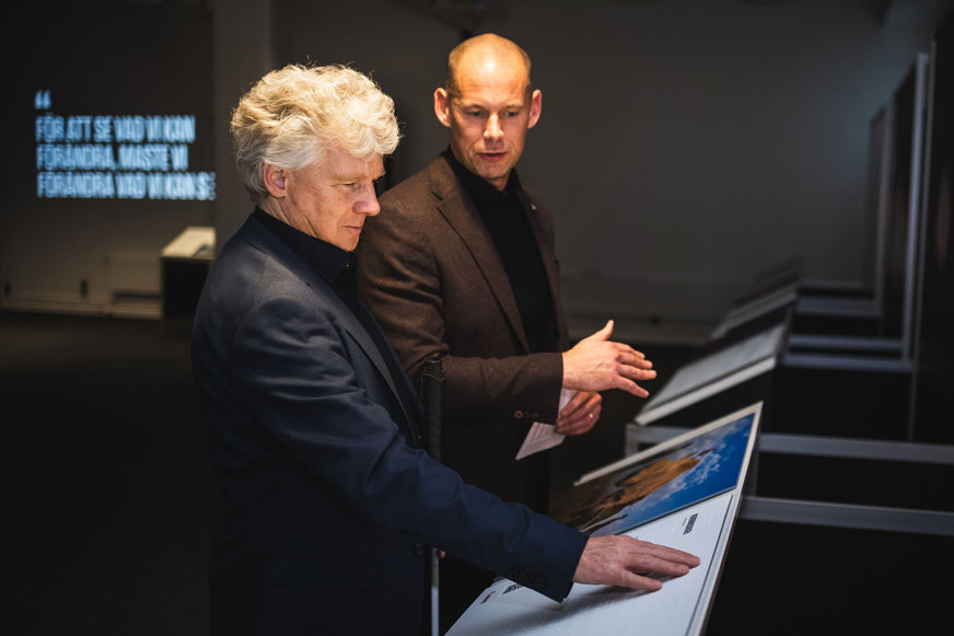 Niklas och Peter står framför fotopanelen med en noshörning. Niklas känner på punktskriftstexten på panelen. Han är klädd i mörk kavaj och har lockigt grått hår. Peter är tunnhårig och är klädd i en brun kavaj. 