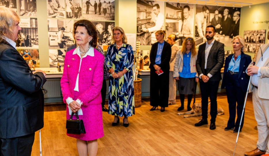 Bild från Synskadades Museum. På väggarna syns fotografier. Drottning Silvia står i mitten i en rosa dräkt och håller en svart handväska. Till vänster står Håkan Thomsson och håller i sin vita käpp. Han är långhårig och bär mörk kostym. Till höger står Linus Forsberg i ljus kostym med vit käpp. Bakom dem står flera personer. En ledarhund ligger på golvet.