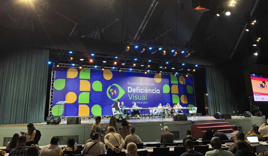 En scen med en grupp människor som sitter uppradade vid ett bord. I bakgrunden en stor skärm med texten "Encontro Mundial da Deficiencia Visual - Sao Paulo 2025". Framför scenen sitter människor och lyssnar.