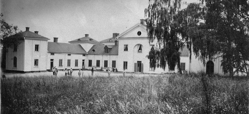 Svartvitt foto på stor vit skolbyggnad, äng och träd i förgrunden.