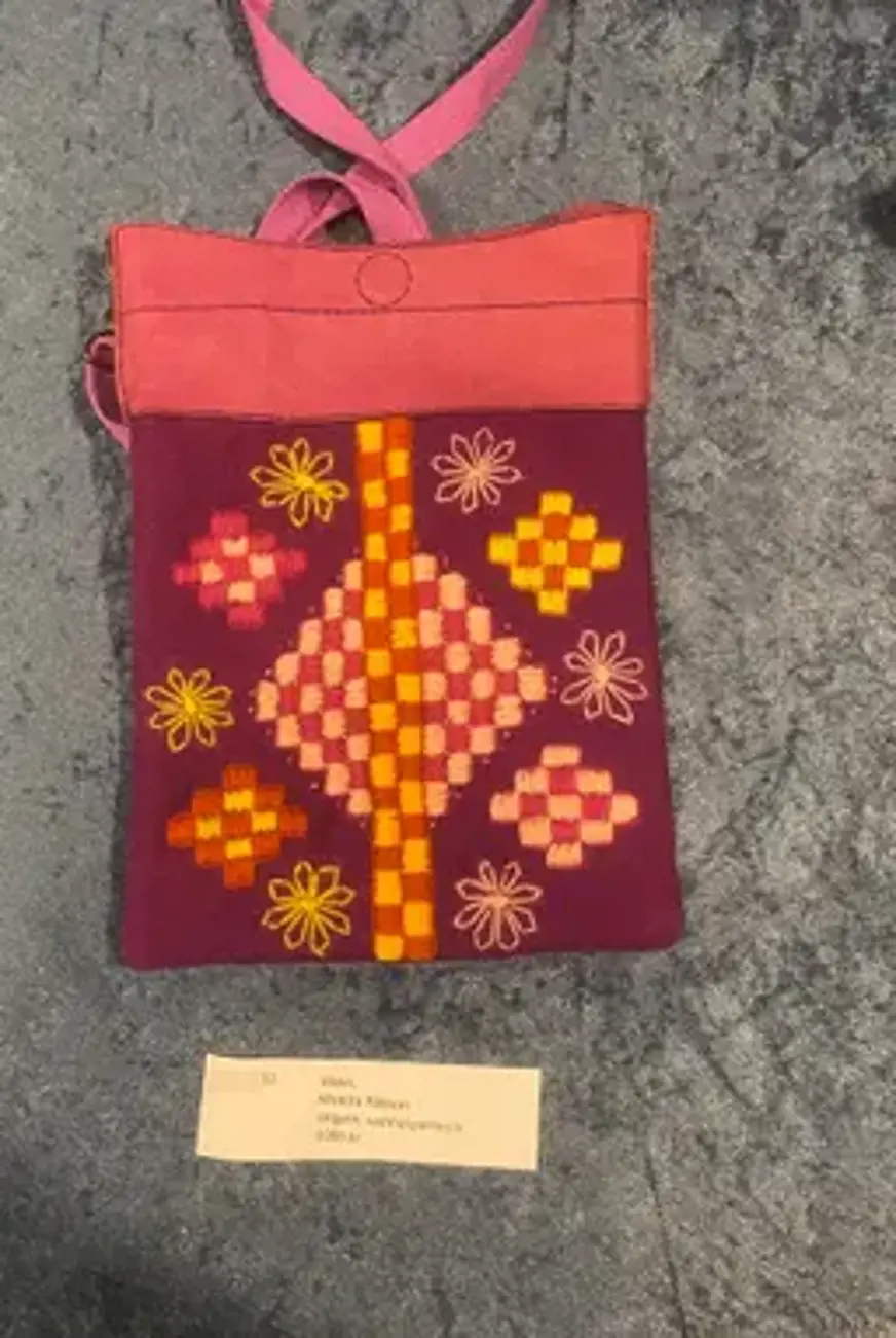 Det är en handväska med en rektangulär form. Väskan är huvudsakligen lila med en rosa övre kant och ett rosa handtag. Den är dekorerad med broderade mönster i gula, orange och rosa färger, inklusive blommor och geometriska former. Det finns en etikett nedanför väskan som säger:  12. Väska, Monica Pålsson Ullgarn, vadmal, pärlor, lin