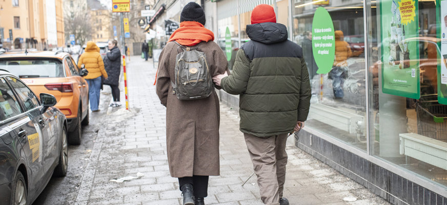Två personer fotade bakifrån på en stadsgata med bilar, skyltar och människor. Den ena personen, med svart mössa, kappa och orange halsduk, ledsagar en person med vit käpp, grön jacka och orange mössa. Trottoaren ser fuktig ut.