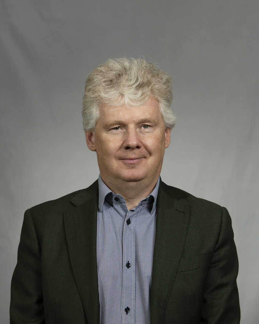 Niklas Mattsson med svart kavaj över smårutig skjorta i blått och vitt. Han har en neutral min och står mot en grå backdrop.