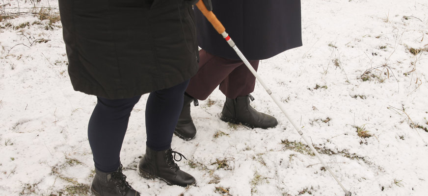 Två personer går på snötäckt mark, en med vit käpp.