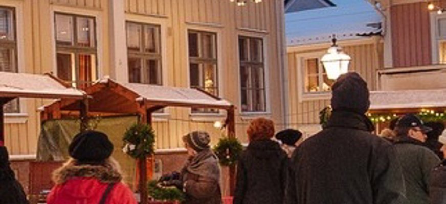 Bilden visar en julmarknad med människor som promenerar mellan små stånd. Ståndens tak är dekorerade med julkransar, och ovanför syns ljusslingor som hänger mellan byggnaderna. Husen i bakgrunden är målade i ljusa färger, och marken är täckt av snö. Stämningen är vintrig och festlig.