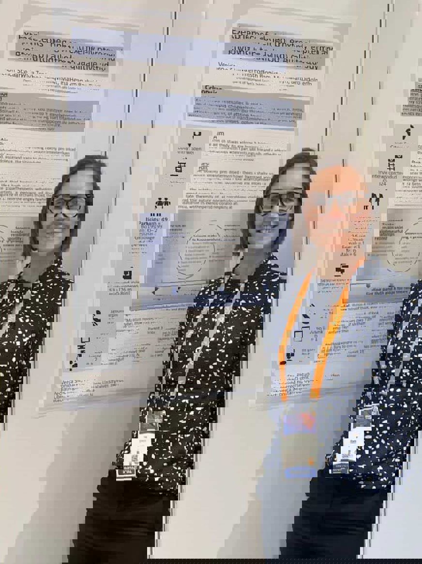 Veronica Starke i kort, brunt hår iklädd svart- och vitmönstrad skjorta. Hon står framför en informationsskylt om erfarenheter av extremt prematura barn.