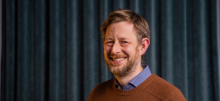 Neil Ormerod har mörkblont hår och skägg och har på sig en finstickad tröja där skjortkragen sticker upp. Han ler med tänderna mot kameran.