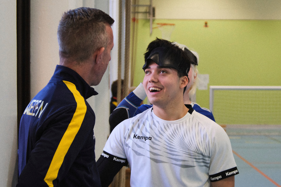 En ung vit man i en vit träningströja pratar med en medelålders vit man i träningsoverallsjacka. Den unge mannen ler och har en svart ögonmask uppdragen i pannan. Platsen är en gymnastikhall och i bakgrunden syns ett goalball-mål