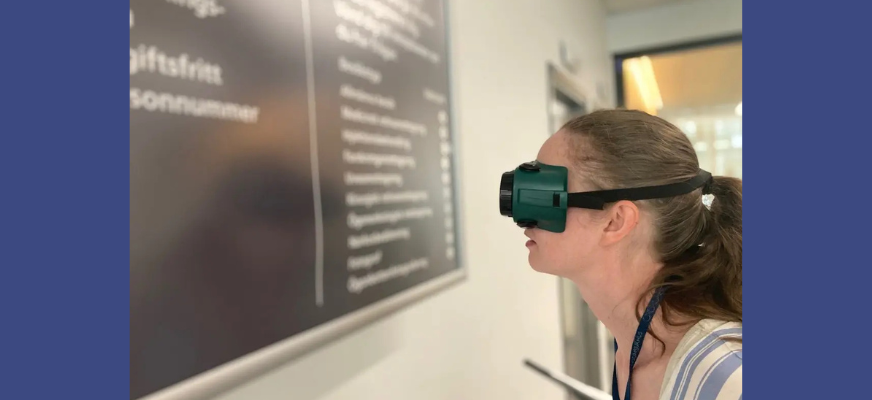 En kvinna med långt brunt hår uppsatt i hästsvans, har fingerad optik för ögonen. Hon står inomhus, framför en skylt med text och provar hur det är läsa med begränsat synfält.