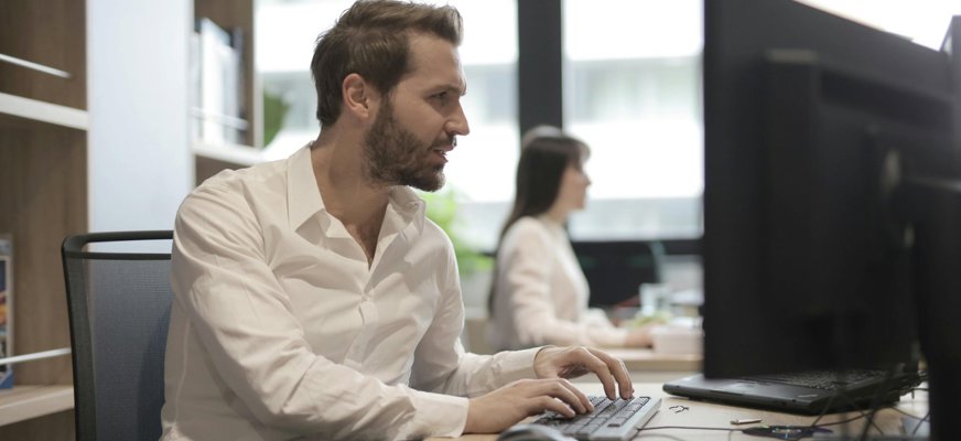 En man i vit skjorta sitter vid ett skrivbord och skriver på ett tangentbord framför en datorskärm. I bakgrunden sitter en kvinna vid ett annat skrivbord och arbetar vid sin dator. Rummet är ljust med stora fönster.