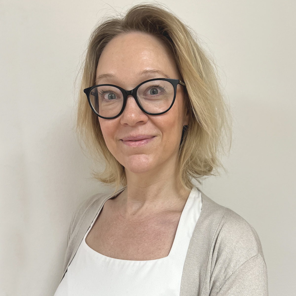 Leende kvinna i 40-årsåldern, stora glasögon med svart skalm, blont axellångt hår, vit t-shirt och beige kofta.