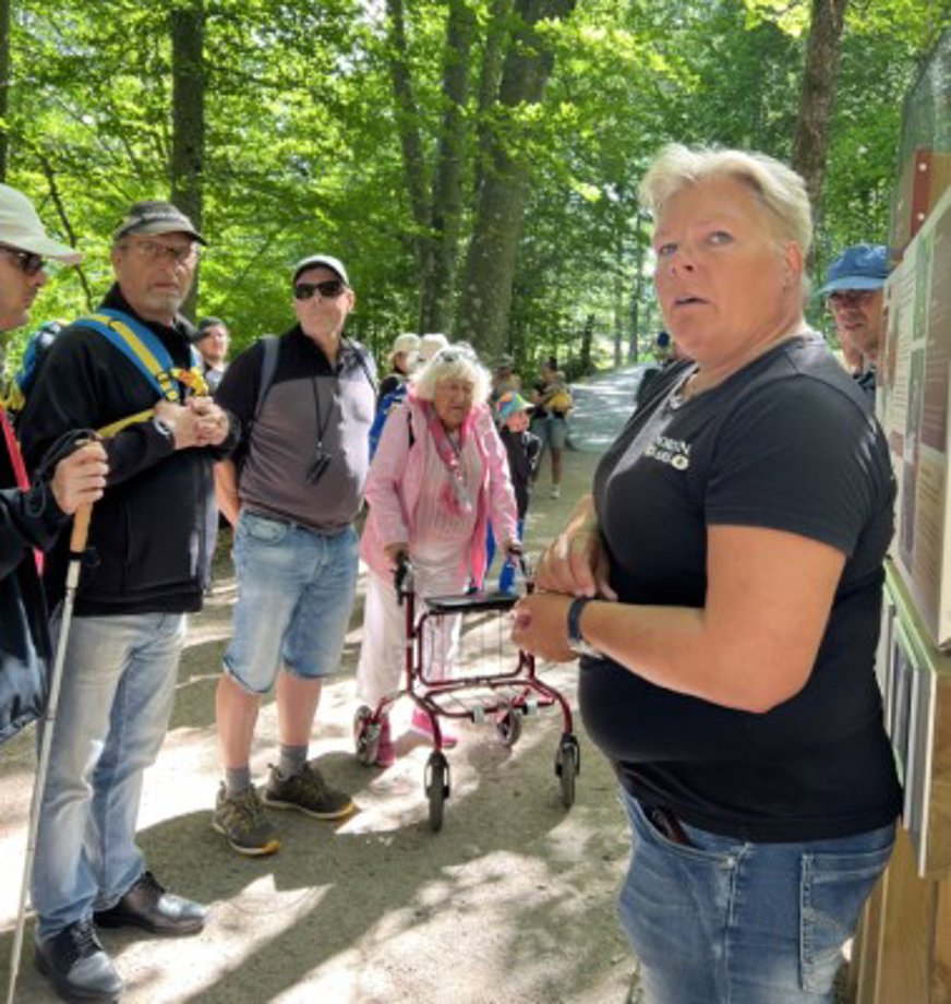 Guiden Eva står och berättar om djuren för en grupp intresserade medlemmar. Det är en solig dag och i bakgrunden syns höga träd med gröna blad.