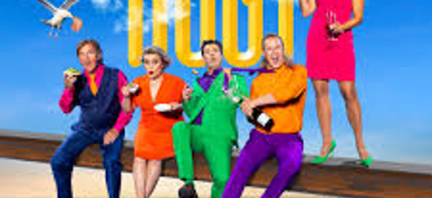 Ensemblen från revyn Högt Spel sitter på en Järnbalk högt upp i luften. 5 personer är klädda i färgglada kostymer.