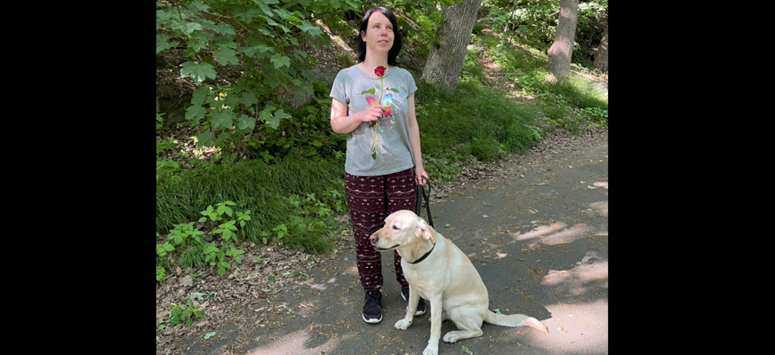 Isabelle står på en väg omgiven av grönska tillsammans med sin ledarhund Smilla, som är en ljus labrador.