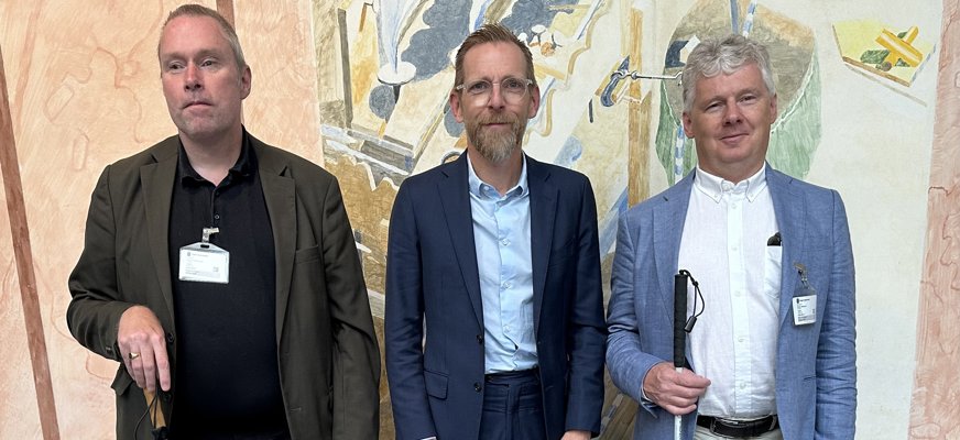 Jakob Forssmed i mitten, blå kostym, glasögon, kort hår och -skägg. På respektive sida representanter från SRF: Två män i udda kavaj, vit käpp. Alla tre uppställda framför kameran.
