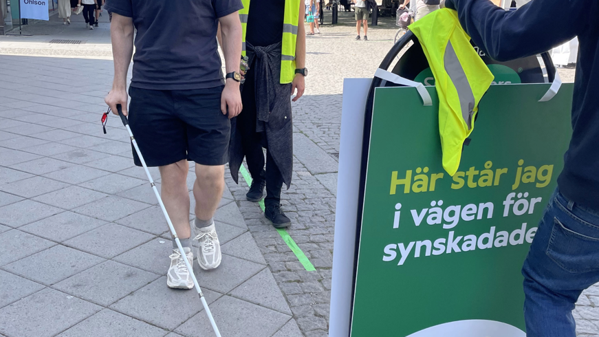 En man med vit käpp promenerar längs den gröna linjen i Visby centrum. Snett bakom honom går en man med grön reflexväst. Framför de båda männen, mitt på den gröna linjen, står en skylt och blockerar. På skylten står det "Här står jag i vägen för synskadade" tillsammans med Specsavers och SRF:s logotyp. Bakom skylten står en person lutad mot skylten.