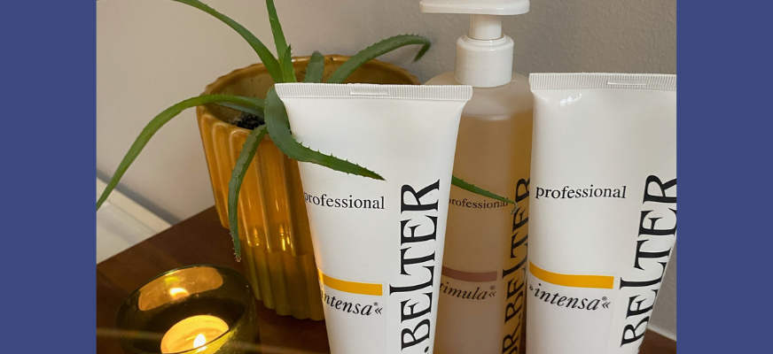 Ett urval hudvårdsprodukter i tuber syns till höger. På vänster sida brinner ett värmeljus i en låg gröntonad glaskopp. Bakom produkterna finns en aloe vera-växt i en senapsgul blomkruka. Bordet är gissningsvis i ek, eller eklaminat. Väggen bakom är målad ljust grå.