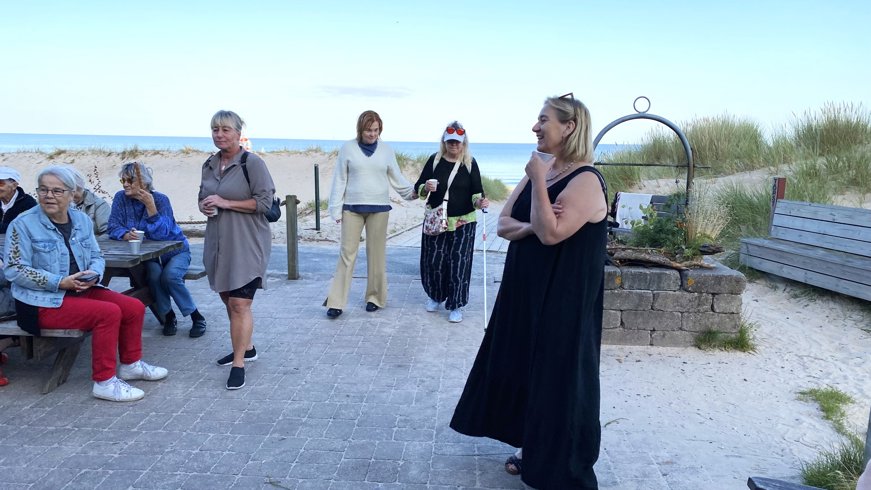 Bilden visar en grupp människor samlade utomhus nära en strand. Några sitter vid ett träbord för picknick medan andra står på ett asfalterat område. I bakgrunden syns sanddyner, gräs och havet. Det ser ut att vara en avslappnad social sammankomst eller möte vid havet. Ingen matematik eller läxrelaterad aktivitet syns på bilden.