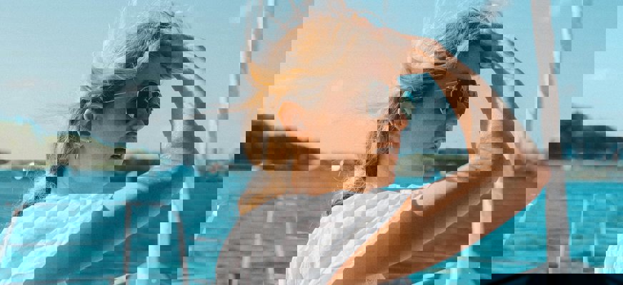 En kvinna står på en båt och blickar långt bort. Hon har ljust år och bär solglasögon. Hon håller i ett vattenglas med den ena handen och för håret bort från ansiktet med den andra.