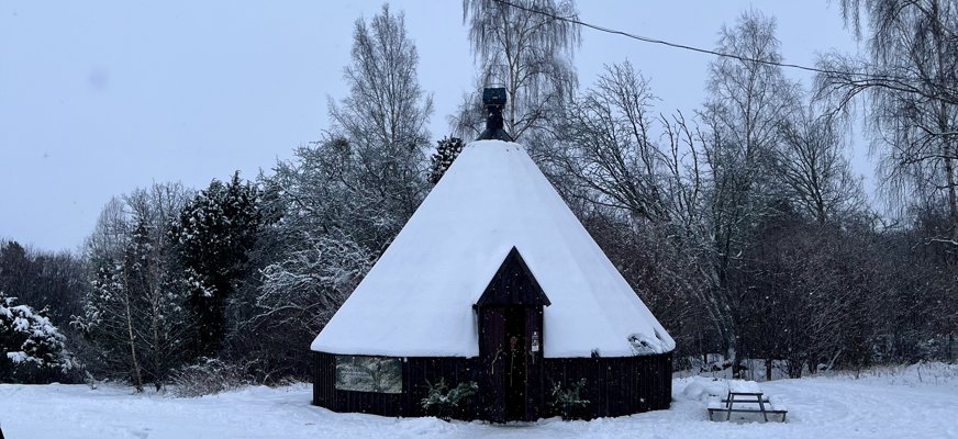 En kåta i ett snöigt vinterlandskap med träd i bakgrunden. En elledning löper tvärs över bilden.