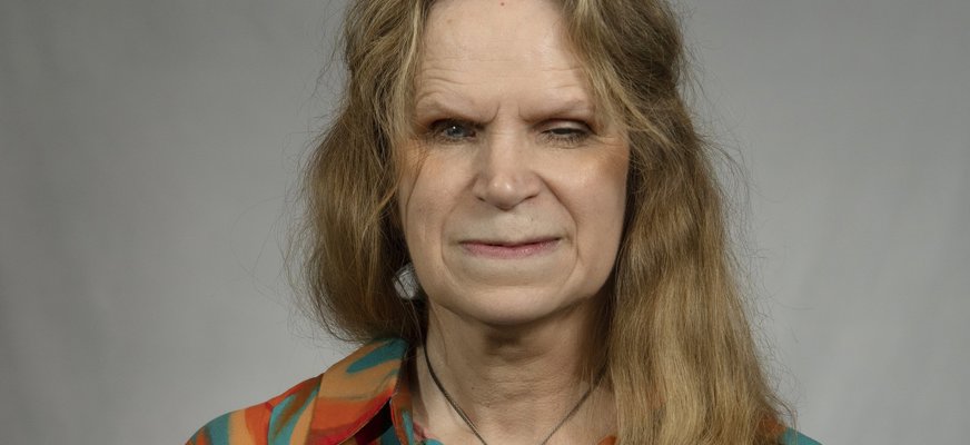 Tiina har långt mörkblont och har på sig en mönstrad skjorta. Hon har ansiktet vänt mot kameran och ler utan att visa tänderna.