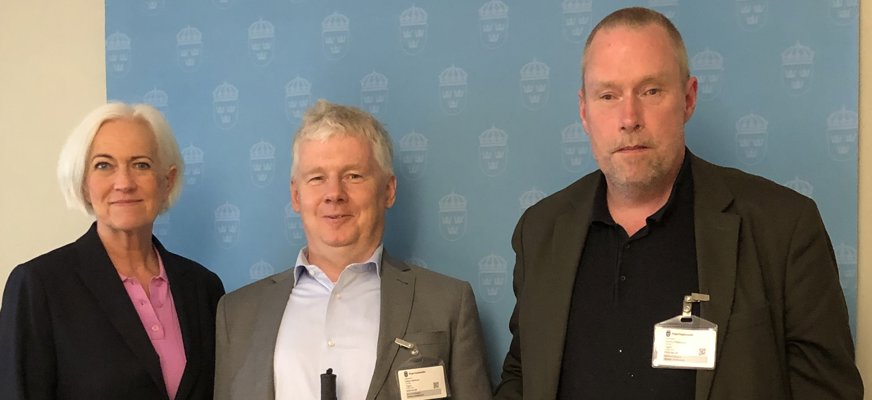 Acko, Niklas och Jimmy står bredvid varandra med en blå vägg med Regeringskansliets logga bakom sig. De har alla tre mörka kavajer och ler litegrann. Niklas och Jimmy håller i sina vita käppar.