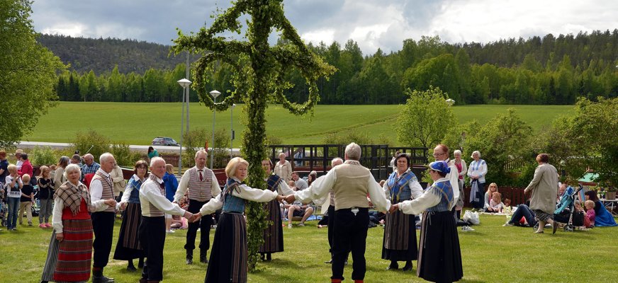 Bilden föreställer en grupp människor i traditionsenliga kläder och folkdräkter som dansar runt en midsommarstång