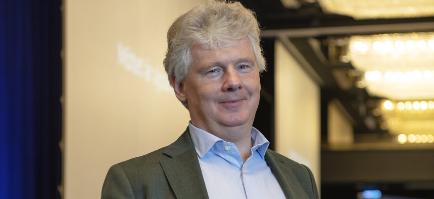 Porträttbild halvkropp, Niklas i ljusblå skjorta, kavaj, chinos, vit käpp. Leendes med kort grått hår.