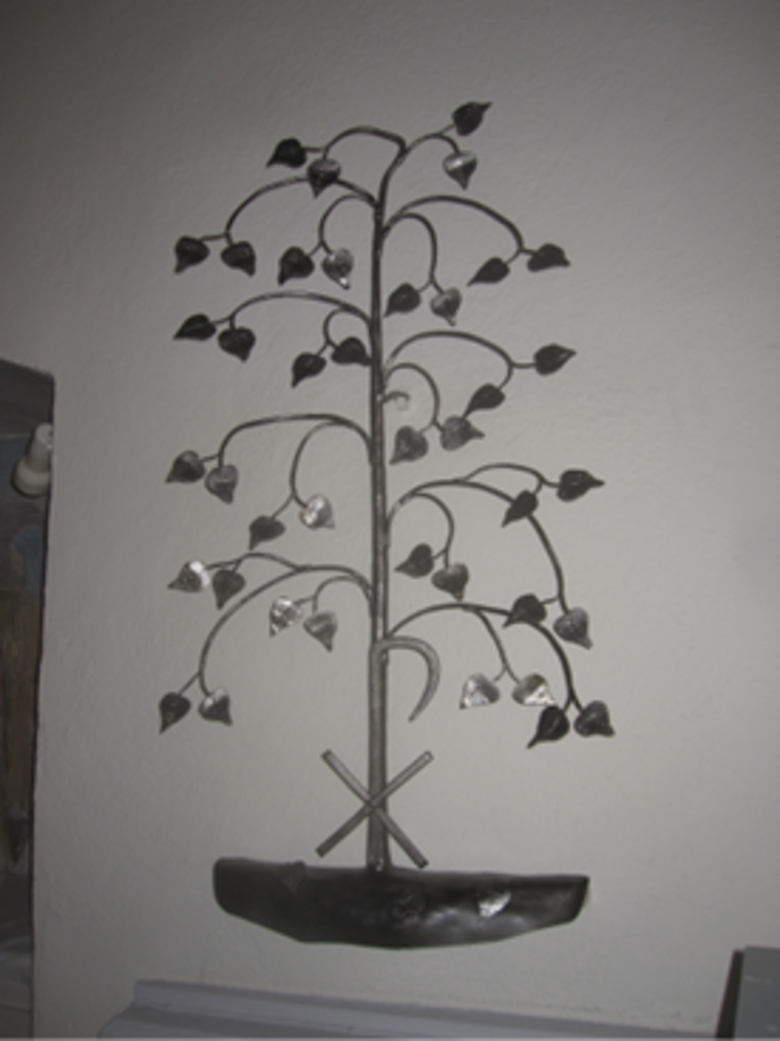 Den metalliska vägskulpturen föreställer ett stiliserat träd med symmetriskt böjda grenar och glänsande blad, vilket ger ett intryck av liv och tillväxt. Ljuset som faller på de reflekterande ytorna skapar ett spel mellan skuggor och blänk, vilket förstärker verkets tredimensionalitet. De korsade verktygen vid trädets fot – en hammare och en skära – kastar tydliga skuggor som ger djup och symbolisk tyngd. Ljuset lyfter fram kontrasten mellan det organiska trädet och de industriella elementen, vilket kan tolkas som en balans mellan natur och arbete. Skulpturen blir därmed inte bara en visuell dekoration, utan en berättelse som förstärks av ljusets rörelse över dess yta.