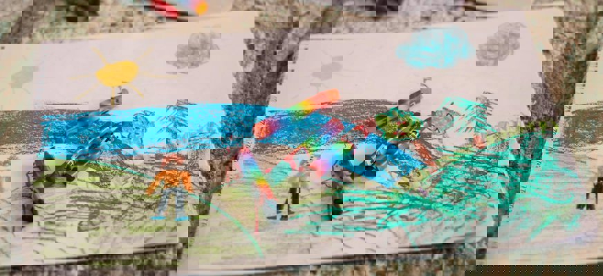 En barnteckning i ett spiralblock ligger på en matta. Färgglada kritor är utspridda ovanpå teckningen som föreställer en sol, vatten, träd och en person i ett landskap.