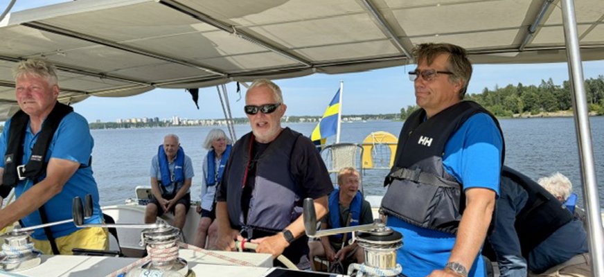 En äldre man med solglasögon styr med båda händerna på båtratten. Han har en besättningsman på var sida om sig. De har blå flytvästar och blå t-shirts på sig. bakom dem sitter en personer i sittbrunnen. I aktern fladdrar en svensk flagga. Delar av storseglet syns ovanför soltaket. Bakom aktern glittrar Mälarens blå böljor i solskenet och vid horisonten syns öar och delar av Västerås.