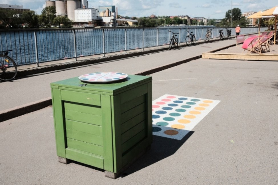 En bred gångväg löper längs vattnet en solig dag. I förgrunden står en grön kubformad låda bredvid ett färgglatt twisterspel målat på marken, och längre bort syns cyklar, solstolar och en uteservering vid kajkanten.