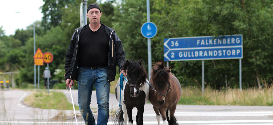 Jan Johannesson går över ett övergångsställe. I ena handen har han en vit käpp. I den andra håller han sina två små hästar.