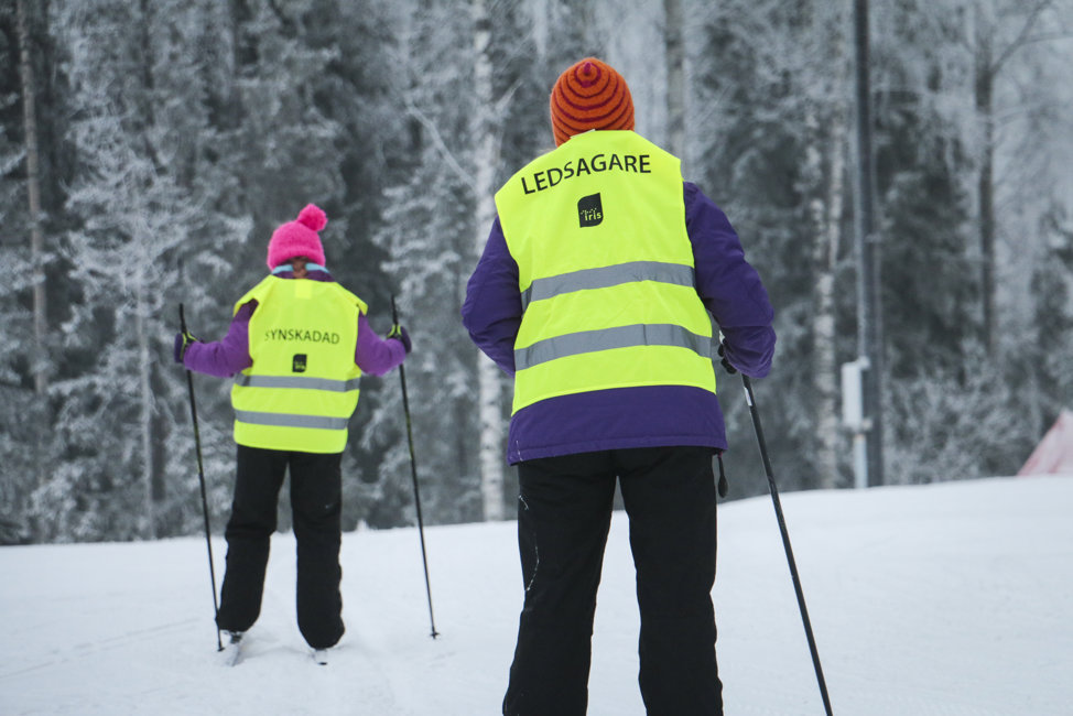 Två personer med varselväst åker skidor, på ryggen står det "ledsagare" och "synskadad"