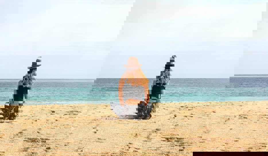 En kvinna klädd i svala kläder sitter i skräddarställning på en solig strand och blickar ut över havet.