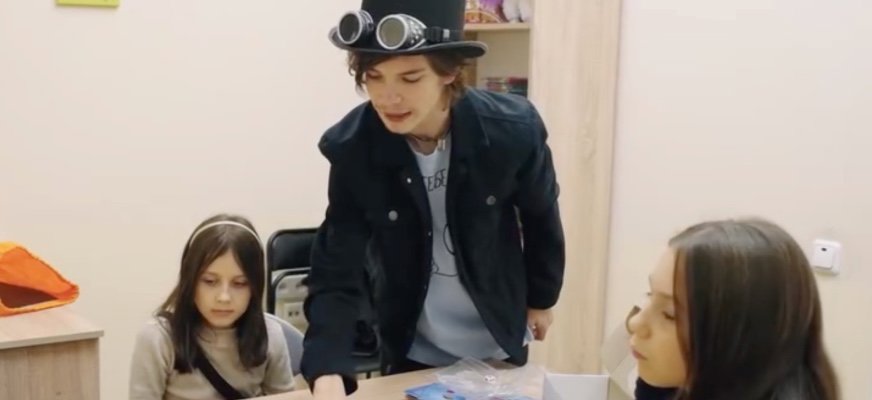 Yaroslav, i hatt med fastsatta glasögon, lutar sig fram över ett bord och visar något. Två barn sitter med vid bordet i ett rum som liknar ett klassrum.