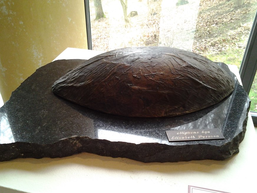 Skulpturen "ellipsens öga" av Elisabeth Persson visar en stor, mörk elliptisk form med grov textur, placerad på en polerad, oregelbunden stenbas. Den är utställd inomhus nära ett fönster, där dagsljus framhäver kontrasten mellan den råa ytan och den släta stenen. Verket för tankarna till naturens former och människans blick – ett öga som betraktar eller blir betraktat. Kombinationen av organiskt och bearbetat material skapar en känsla av stillhet och närvaro.