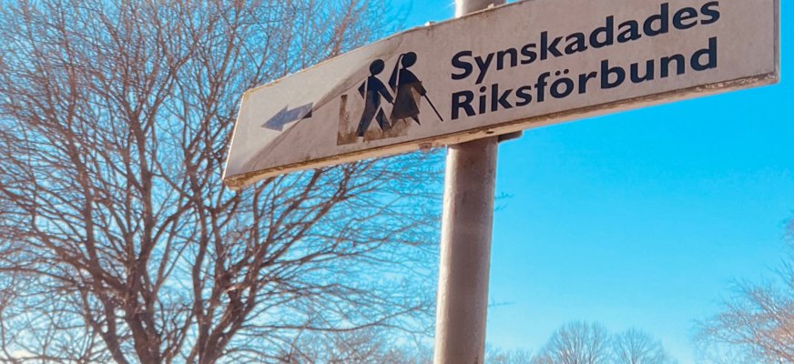 Bild på en vägskylt med texten "Synskadades Riksförbund", framför en pil. Bakom en blå himmel och träd med kala grenar.