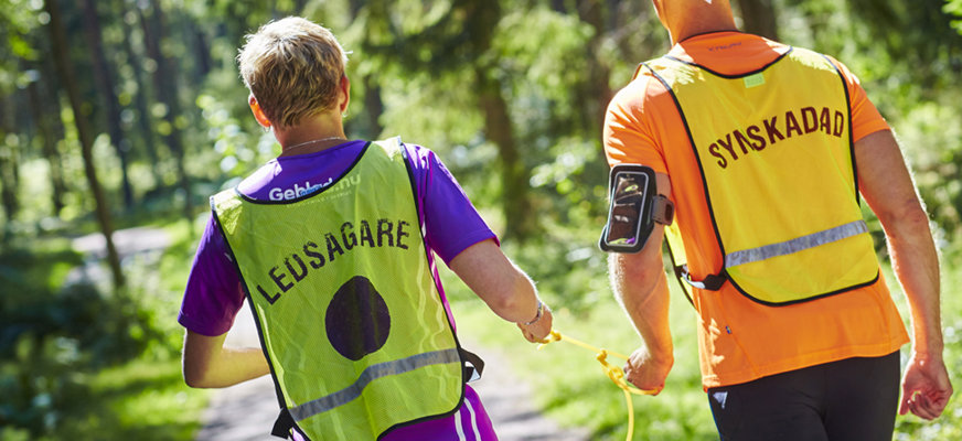 Två män som joggar tillsammans på en skogsväg. De har varsin reflexväst med "Ledsagare" respektive "Synskadad" textat på ryggen.