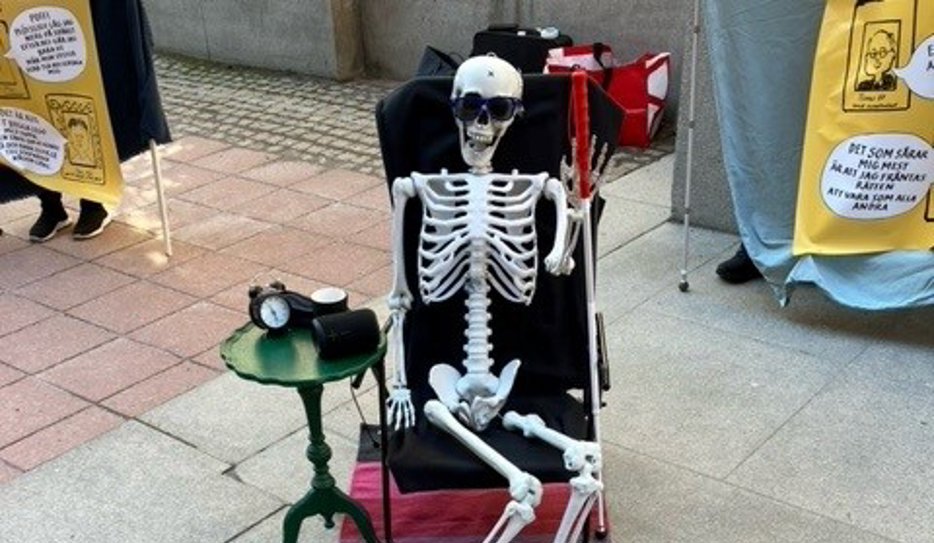 Ett låtsasskelett sitter på en svart solstol med svarta solglasögon och handen uppe i en vinkning. Bredvid skelettet står ett grönt nattduksbord och under en matta med röda, lila, rosa och svarta ränder. 