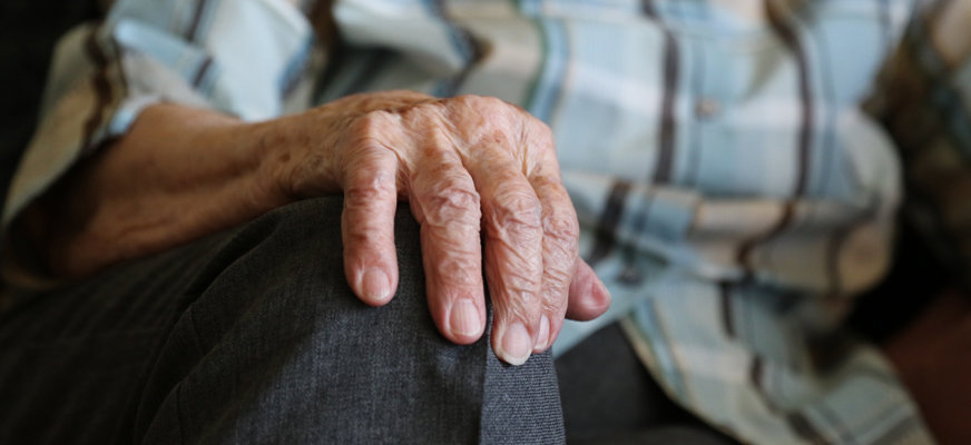 En äldre mans hand vilar mot hans gråklädda knä. I bakgrunden syns mannens rutiga skjorta i blått, vitt och beige.
