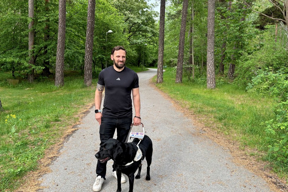 En atletisk man i 30 års åldern står på en skogsväg med sin svarta ledarhund i sele. Han har kort brunt hår, svart t-shirt, svarta jeans och vita sneakers.