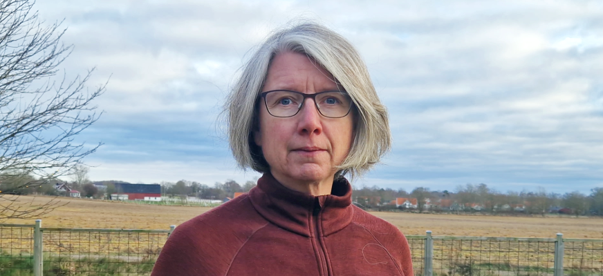 En kvinna står framför en äng i ett höstigt landskap. Hon har kort grått hår klippt i page, en röd funktionströja och gladögon med svarta bågar. Kvinnan ser med allvarlig blick in i kameran.