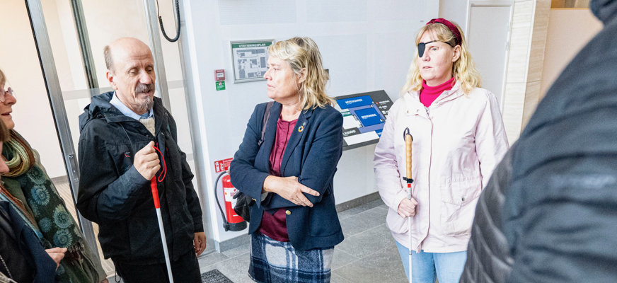 Henrik Eldh håller sin vita käpp till vänster i bild. Birthe Sandberg står i mitten av bilden med armarna i kors över bröstet. Hon har en mörkblå kavaj, rutig kjol och en handväska över högeraxeln. Bakom henne står Helene Jangenfält och lyssnar på samtalet. I bakgrunden syns en taktil karta över Rådhus Skånes lokaler.